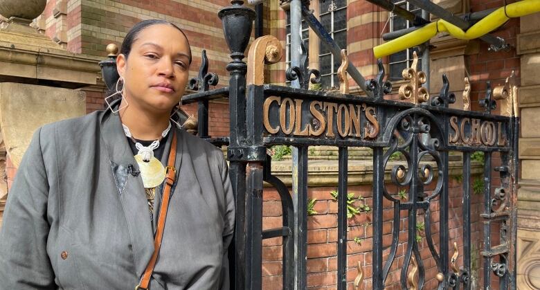 A woman with black hair, wearing a grey coat, stands beside a wrought-iron gate with a sign that reads 'Colston's School.'