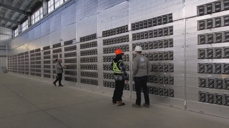 A massive grey wall in a warehouse willed with bitcoin miners. Three people, two wearing hardhats, stand in front of the wall.