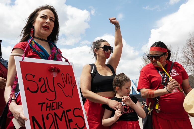 Three adults in red stand in the frame as well as a young child. The adult on the left holds a red sign that says, 