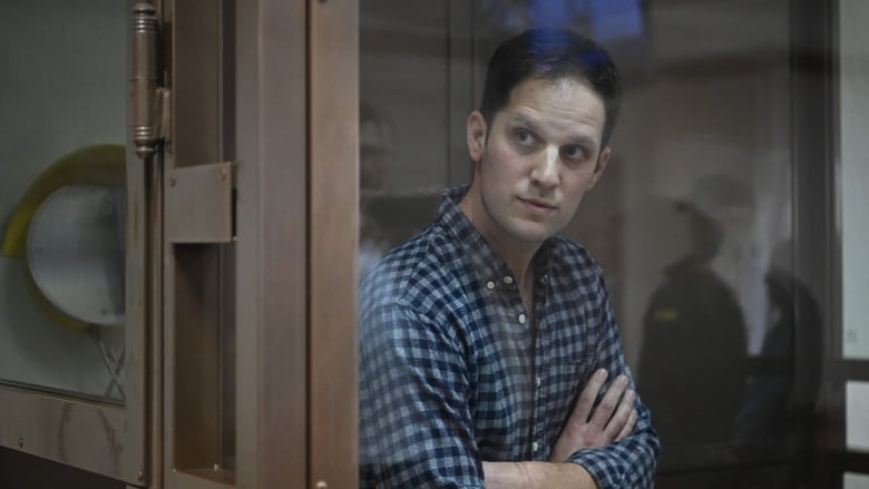 A man wearing a blue plaid shirt stands in a glass box in a courtroom, with his arms crossed in front of his chest.