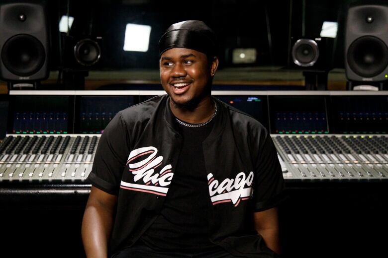 A smiling man with a wearing a durag and black Chicago baseball jersey sits in front of a studio control board.