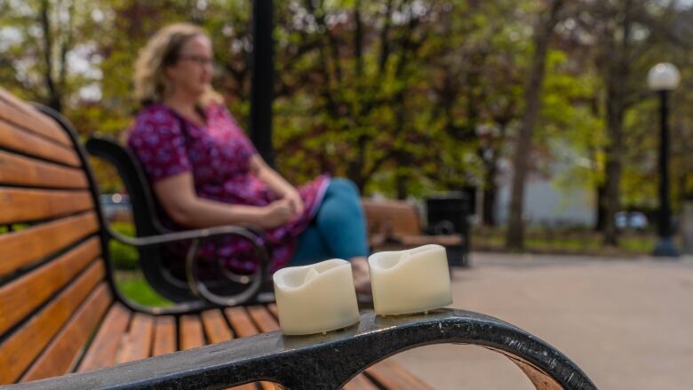 Two candles sit on a park bench arm rest. A woman sits on the park bench, blurred in the distance.