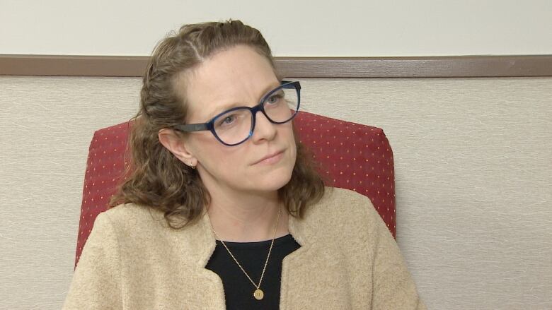 A woman with brown hair, blue framed glasses and a gold necklace looks off camera.