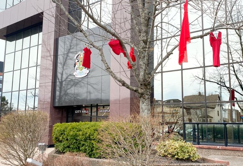 Five red dresses hang from the branches of a tree outside the front of a building labelled city hall.