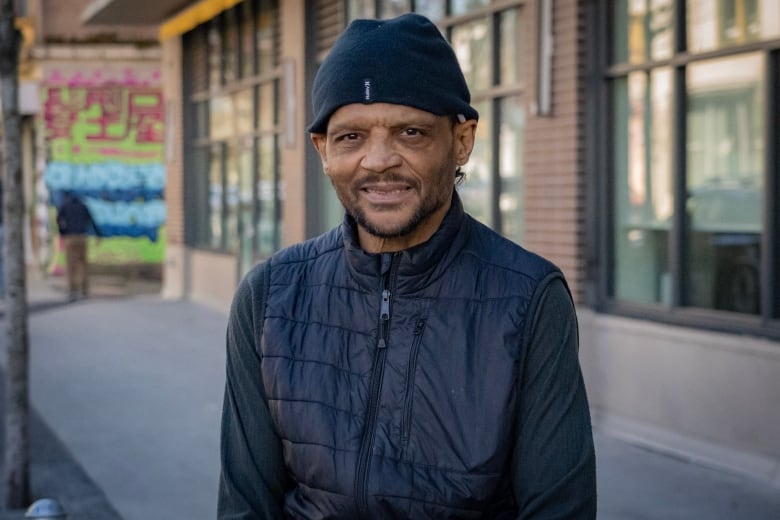 a man wearing a black sweater, black vest and black toque smiles in front of a brick building