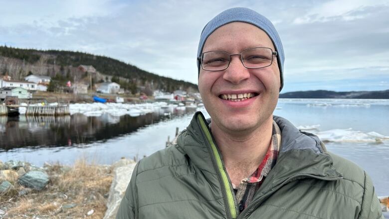 A man in a blue toque and green winter coat smiles at the camera, against an ocean outport background.