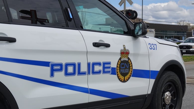 A white SUV with diagonal blue stripes has a CN Police logo painted on its side. The logo shows the words CN Police surrounded by golden laurels and topped with a crown.