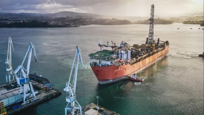  A 300-metre rusty ship being assisted by tugboats leading it into a port with cranes. 