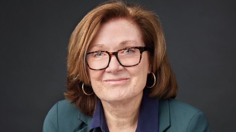 A portrait of a woman with dark hair, glasses and a dark green blazer smiles at the camera.