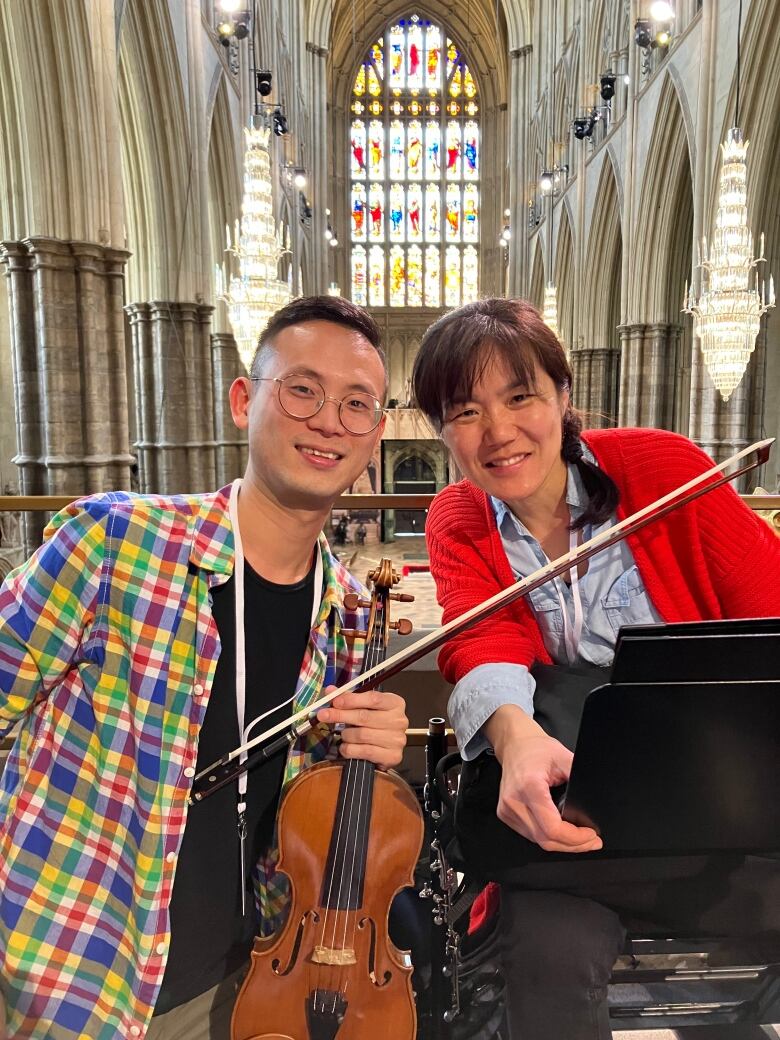 A man holding a violin and a woman behind a music stand smile in front of a stained glass window in the Westminster Abbey