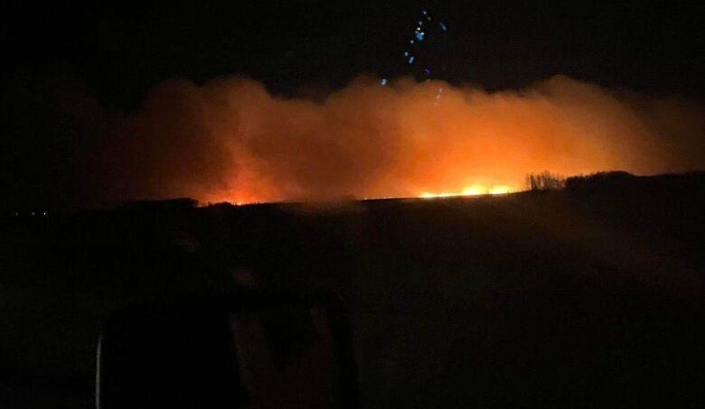 Clouds of smoke glow orange in the nights sky as a wildfire burns on a wooded hill.