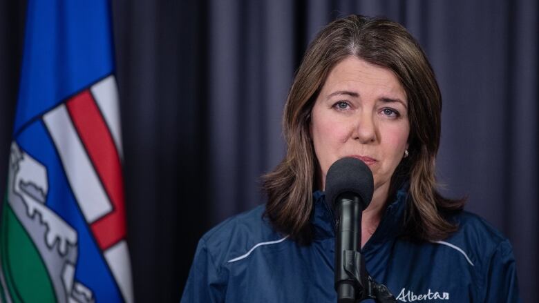 A white woman with brown hair down to her shoulders is wearing a navy blue windbreaker. She is standing in front of a microphone and next to an Alberta flag.