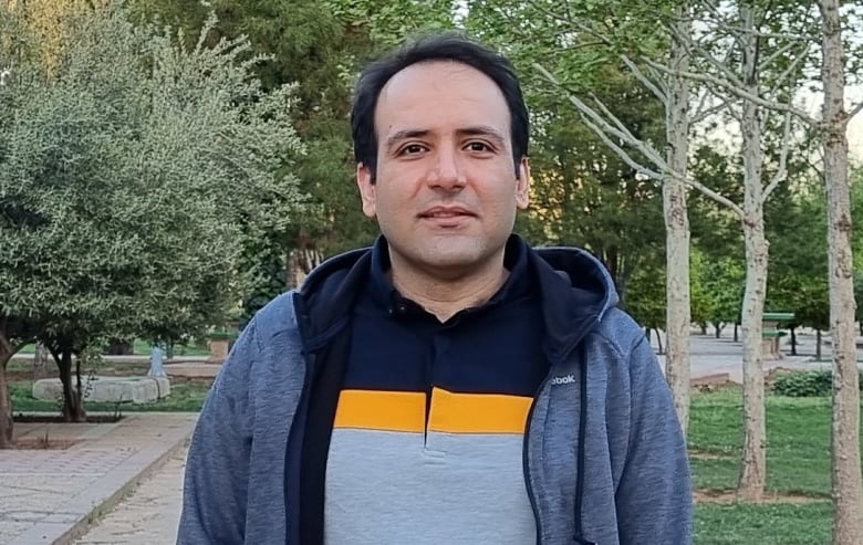 A man in a polo shirt and jacket stands outside, smiling at the camera.