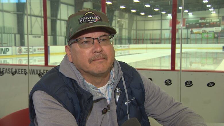 A man in a hoodie, vest and baseball hat sits in front of an indoor hockey arena being cleaned by a Zamboni.