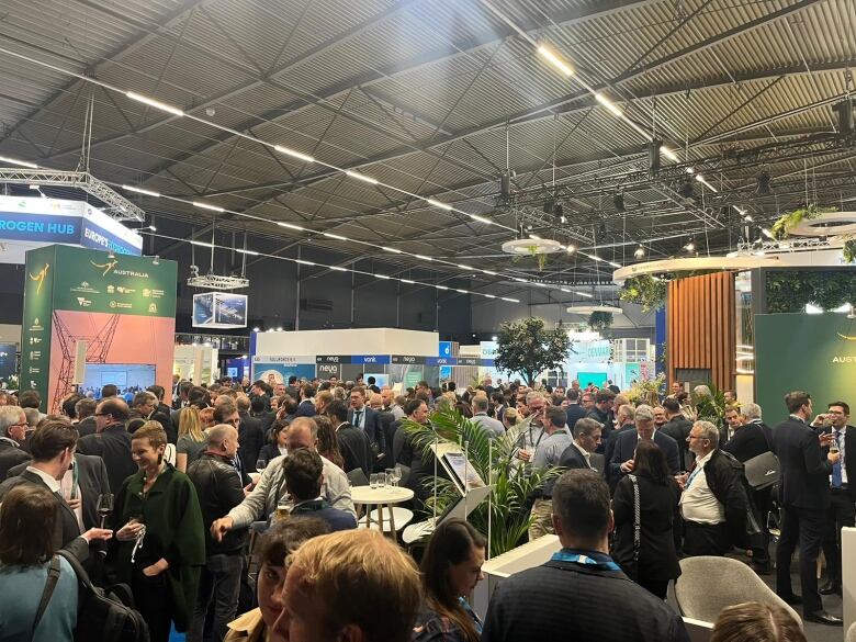 A crowd of people in a large room for an exhibition with booths around the room.