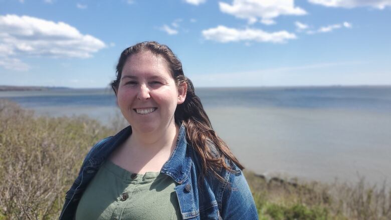 A woman with dark hair wears a jean jacket and green dress while smiles at the camera. She stands near a grassy cliff that overlooks the Bay of Fundy. 