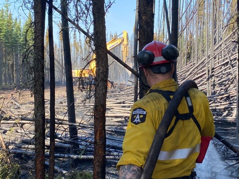 Firefighter in bright yellow clothing and helmet works in treed area with big machinery seen in the distance