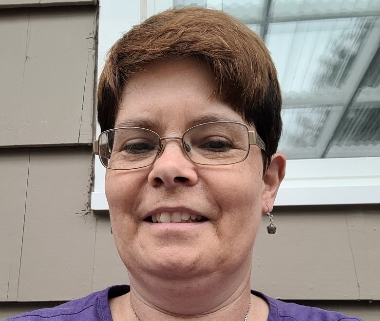 A woman with auburn hair in a purple shirt and glasses smiles for a photo. 