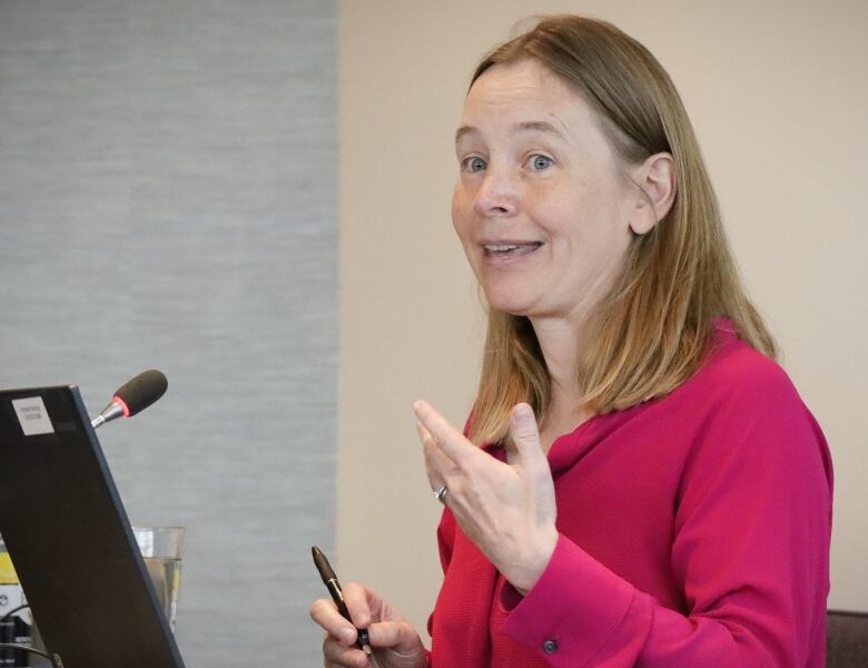 A woman in a pink shirt gestures with her hands as she speaks in front of a microphone.