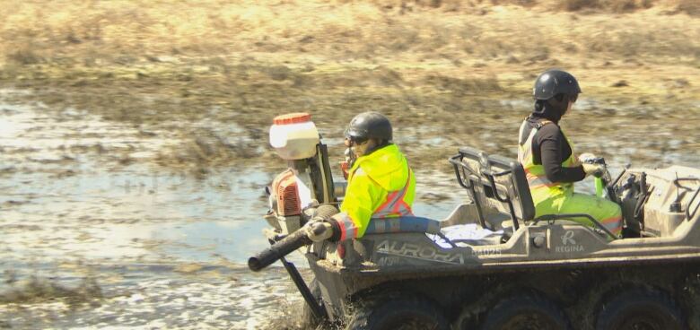 Two city workers riding a four wheeler spray a field with pesticides. 