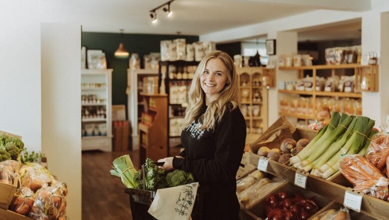 A blonde woman in a black sweater smiles at the camera as she holds a basket of produce. She stands in the middle of a market with shelves of vegetables 