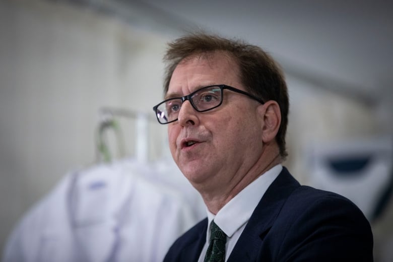 A man wearing glasses speaks in front of lab coats in the background.