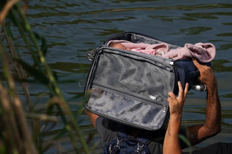 Wading in water, a person holds a suitcase above his head, which is open and carrying a baby inside. 