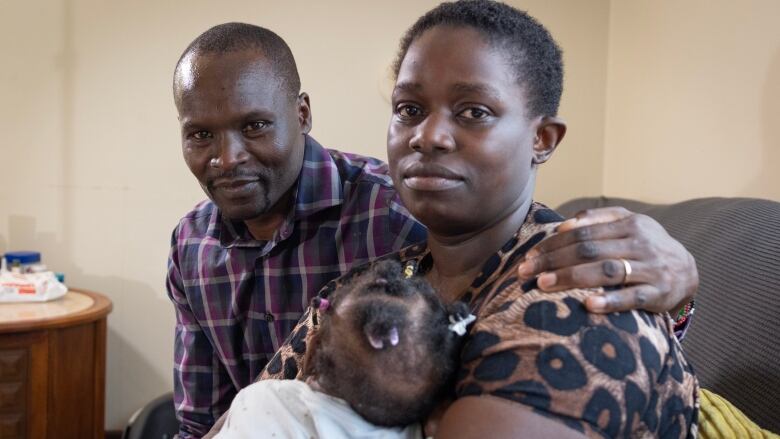 A man sits with his arm around his wife, who holds their baby daughter.