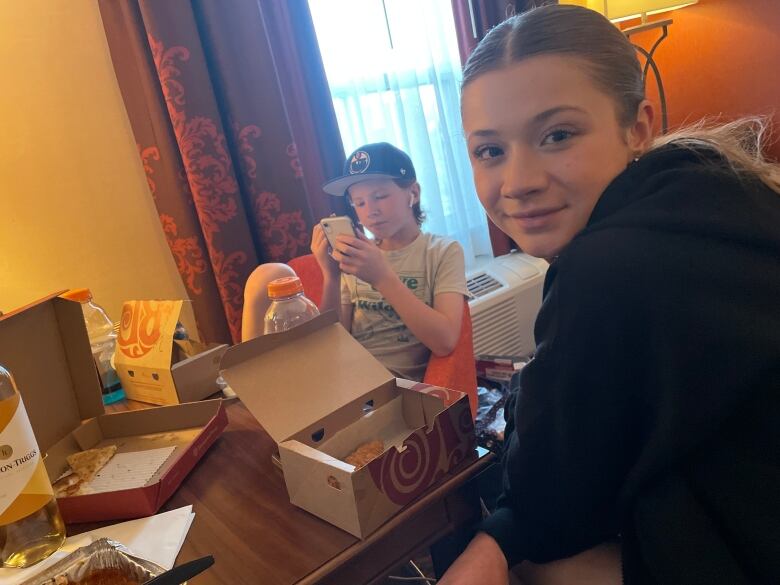 A boy, looking at his phone, and a girl sit at a hotel table with takeout boxes.