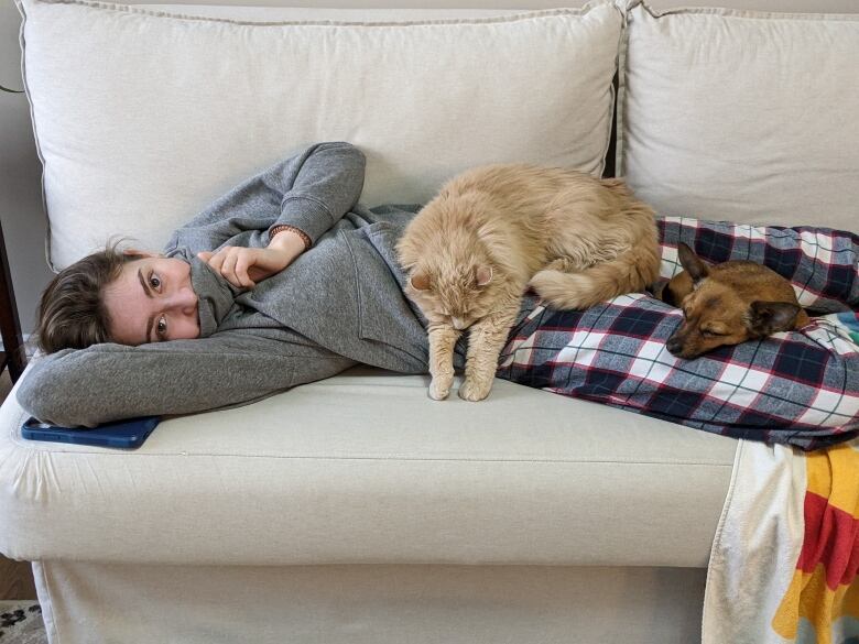 A girl lies on a couch with a dog and cat lying in her lap.