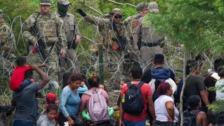 A group of about a dozen people stand on one side of a barbed wire fence. On the other side are uniformed guards.