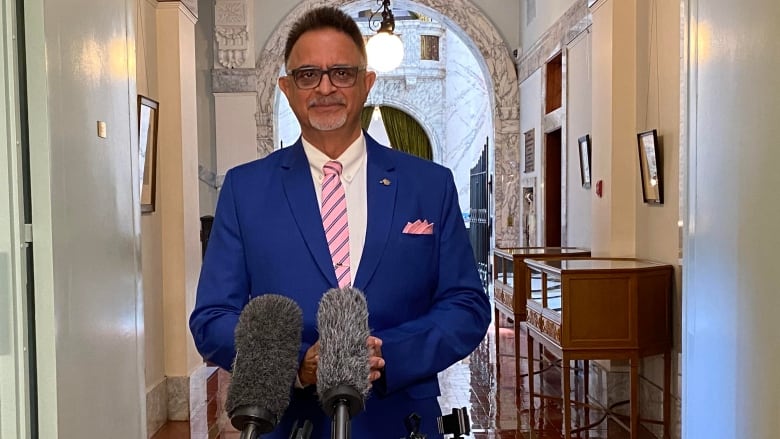 A man in a blue suit and striped pink tie stands in front of a bank of microphones in a B.C. Legislature hallway.