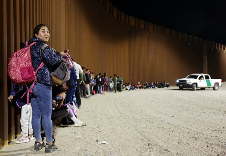 People carrying bags and knapsacks stand in a long line up against a brown wall. 