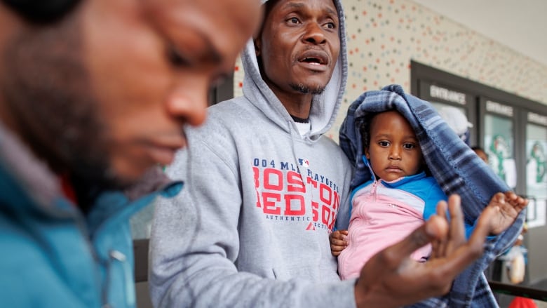 A man in a hoodie gestures with one arm, while holding an infant girl in the other. She is wrapped in a blanket. Another man, his eyes downcast, is in the foreground.