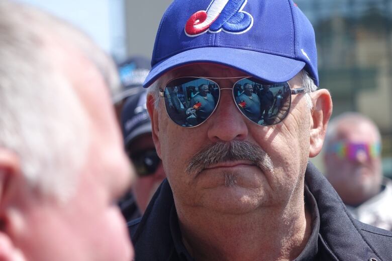 A man wearing sunglasses and a blue baseball hat staring at another man in the foreground. 