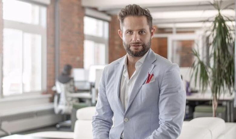 A young bearded man in a light suit and an open-necked white shirt sits on the edge of a white couch in a tastefully decorated apartment, looking directly into the camera with a neutral expression.