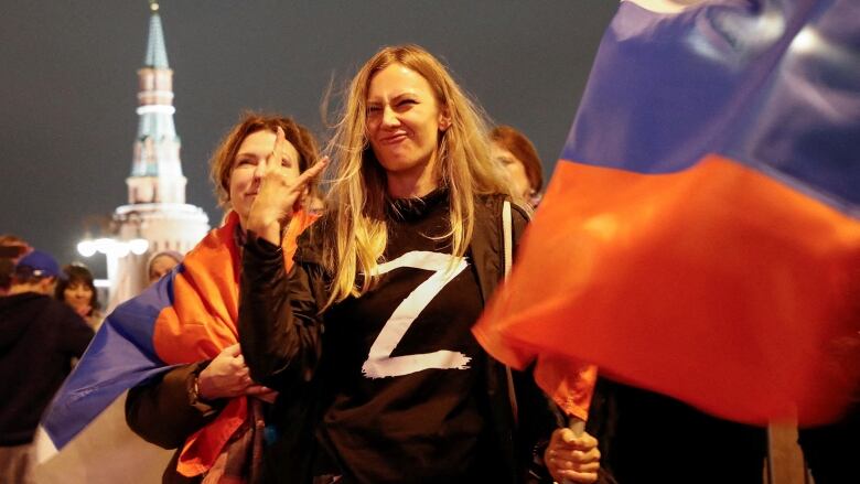 A woman with long blond hair, wearing a black shirt with a white letter Z on the front, makes a hand gesture while holding a red, blue and white flag.