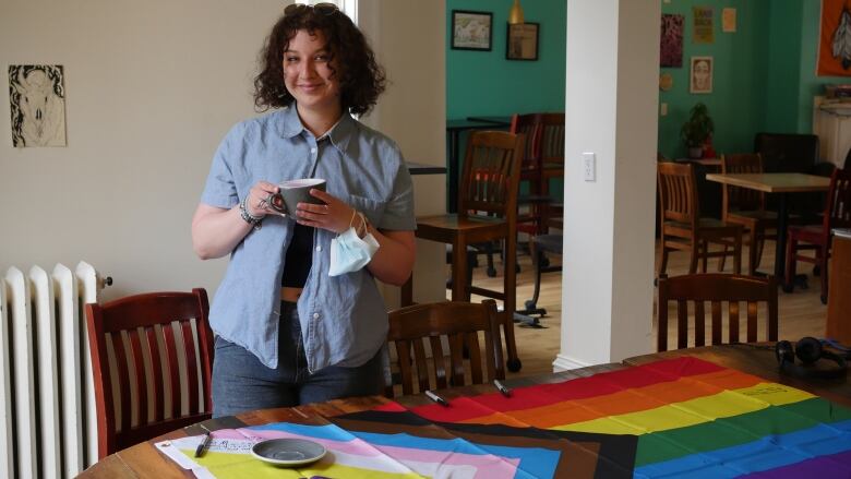 An individual, who is standing up, holds a coffee cup in front of a table. On the table, is a progress pride flag, which in addition to the usual rainbow, includes black and brown stripes to represent communities of color, along with the colors pink, light blue and white, which are used on the transgender pride flag.