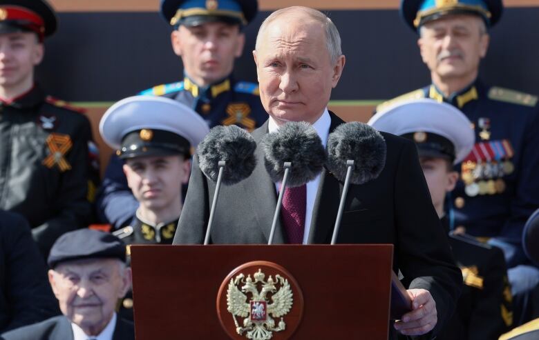 A man with grey hair, wearing a dark suit, speaks into microphones at a podium, as people in military uniforms stand behind him.