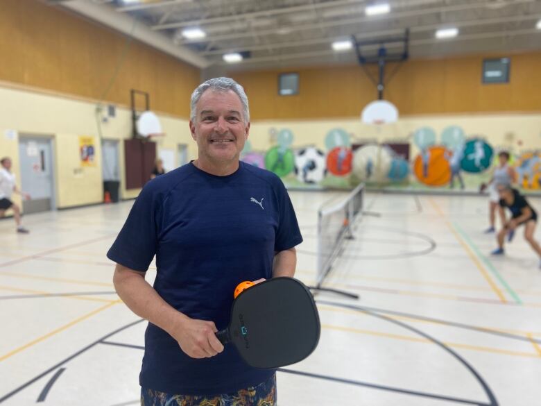 Man in navy shirt standing in front of pickleball court in indoor gym with paddle and orange wiffle ball in his hand, smiling. 