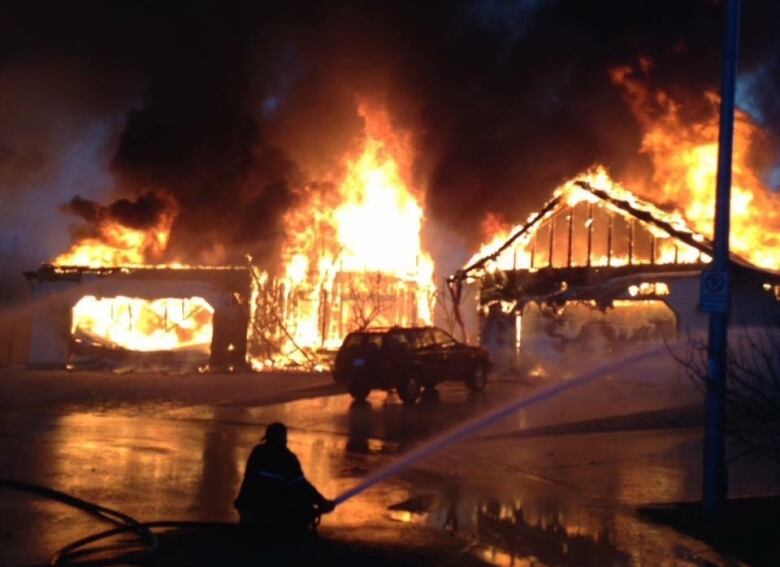 A house is consumed by fire, as a silhouetted figure in the foreground sprays water from a firehose. 