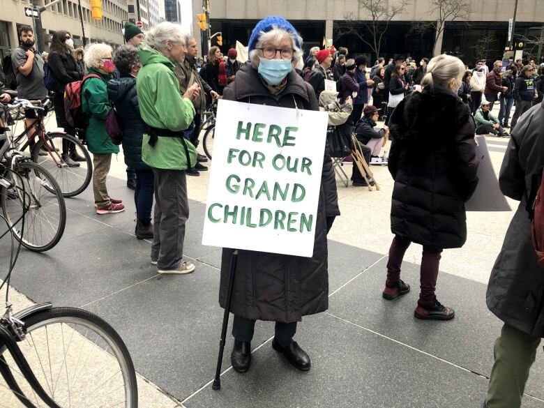 Elderly woman holds up sign that says 'Here for our grandchildren.'