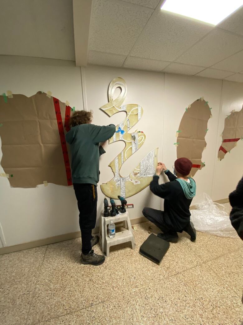 Two people work on a piece of art hung on a white wall.