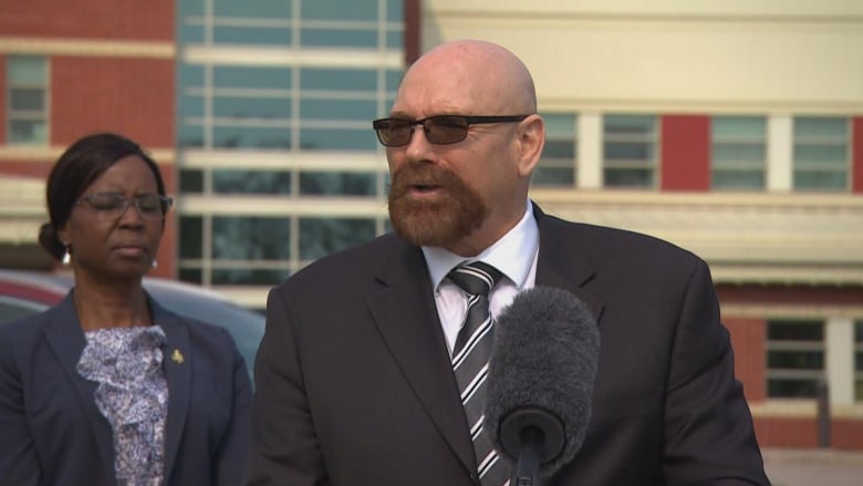 A man in a black blazer, dark stripped tie and white shirt speaks with media.