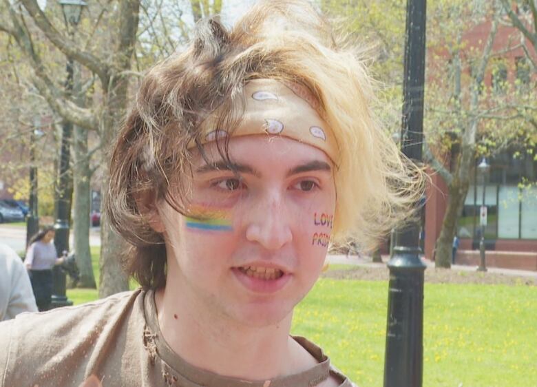 A person with brown and blonde hair with a beige bandana pushing it back. They have a rainbow on their left cheek.