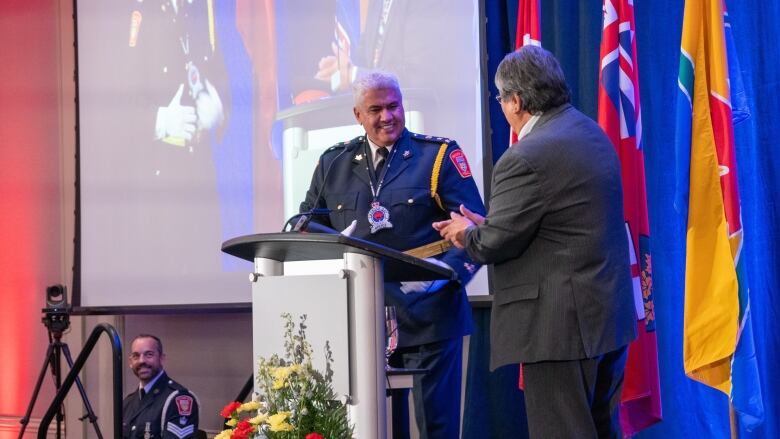 A man in a police uniform smiles while standing at a podium next to a man in a suit.