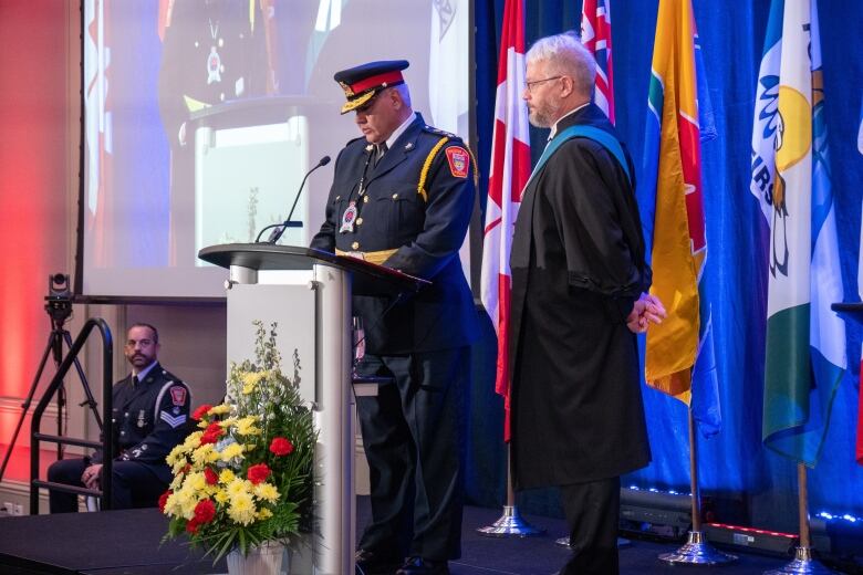 A man in a police uniform reads at a podium while a man wearing justice of the peace robes looks on.