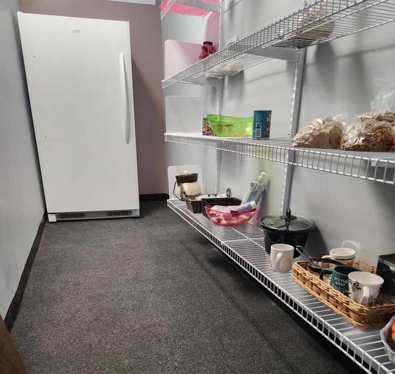 A white refrigerator stands at the back of a thin room that is lined with shelves on the right side.