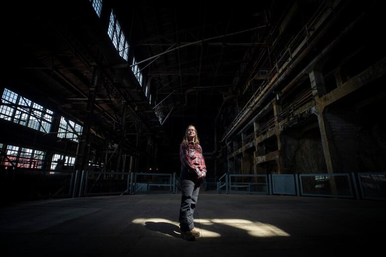 A lady in a plaid shirt looks into the distance in a dark room as natural light shines down on her. 