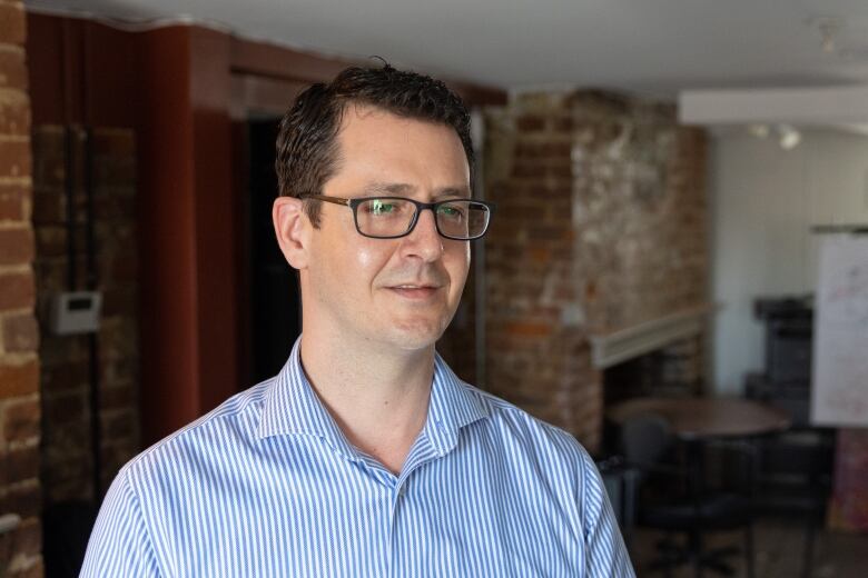 A man wearing glasses is shown wearing a dress shirt with thin blue and white stripes and in the background is a red brick wall inside an office.